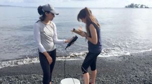 A professor reads off an instrument while her student records information, on the blacksand shore of the bay.