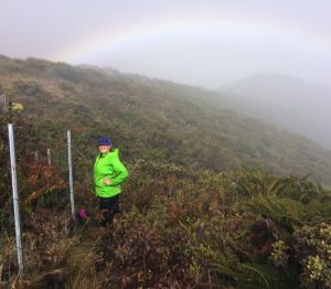 A student stands near a fence on a scrub-covered hillside with a faint rainbow in the mist behind her.