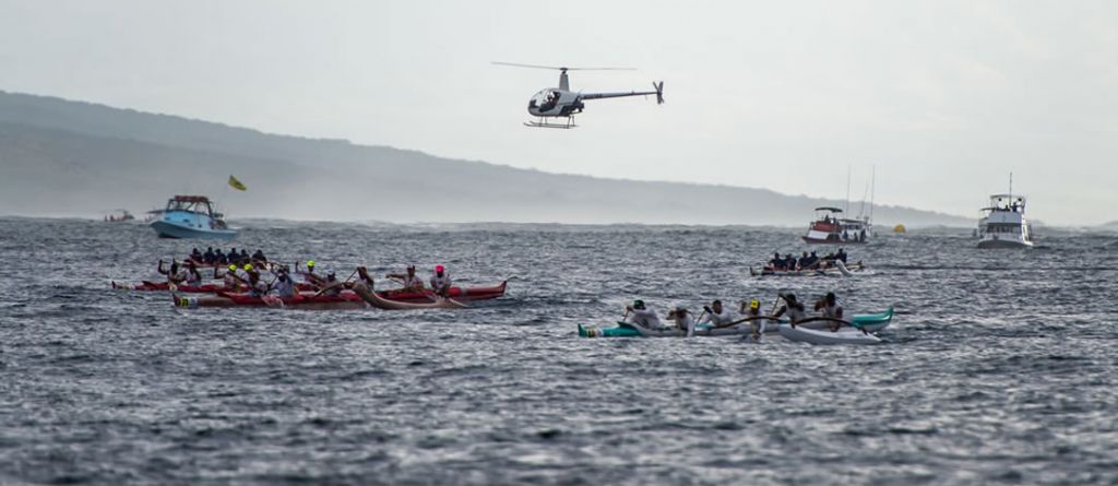 Several canoes and teams racing in ocean with support boats and helicopter surrounding.