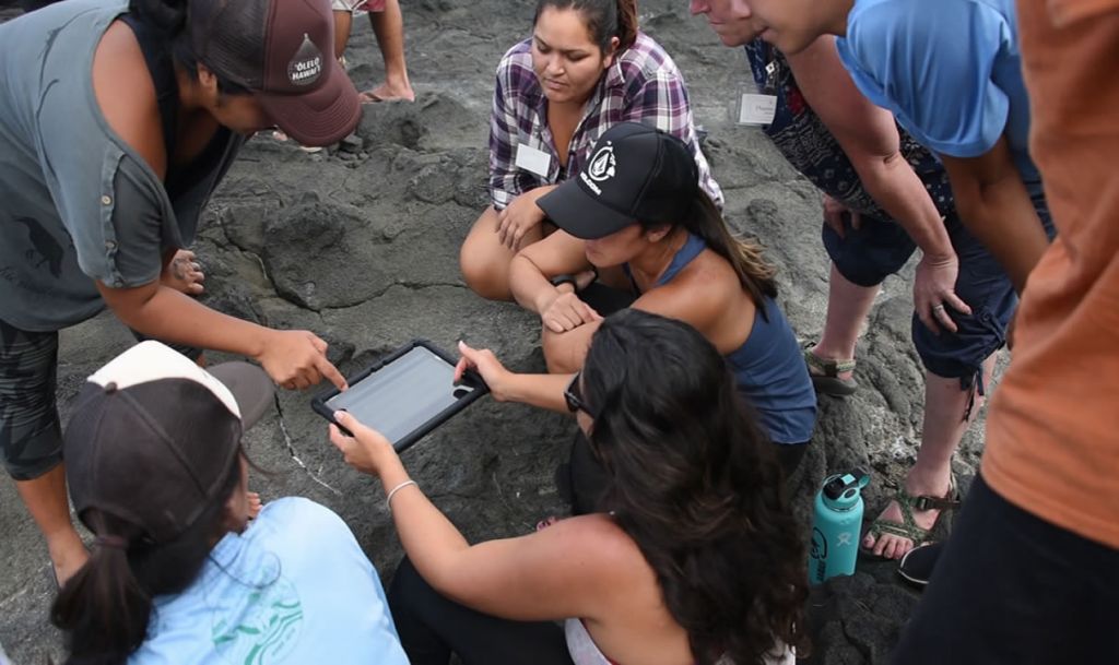 Group of participants on rocky shoreline gathered around tablet and learning.