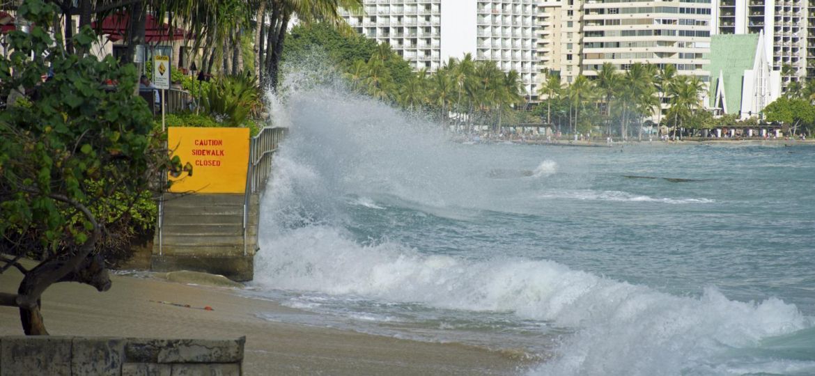 Waves splash against a railed cement wall that bears a "Caution: Sidewalk closed" sign.
