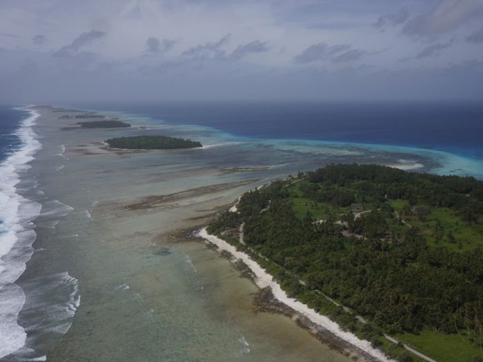 Airplane view looking down along a line of small, tree-covered islands surrounded by shallow lagoons and ocean waves