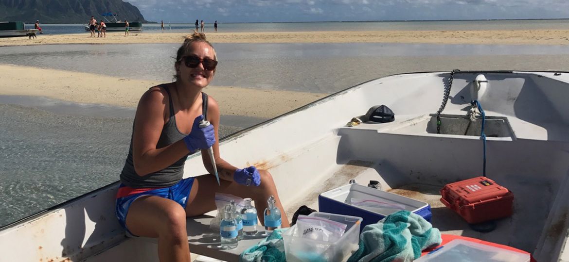 A student, grinning at the camera, sits in a small boat beached on a sandbar while she takes water samples from a bag