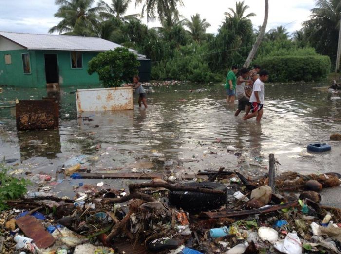 Local children wade through calf-high dirty water, past flood accumulated trash and a flooded house.