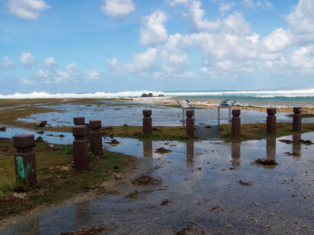 Pilons and interpretive signs border flooded walkways and grassy areas as the surf lies feet away.