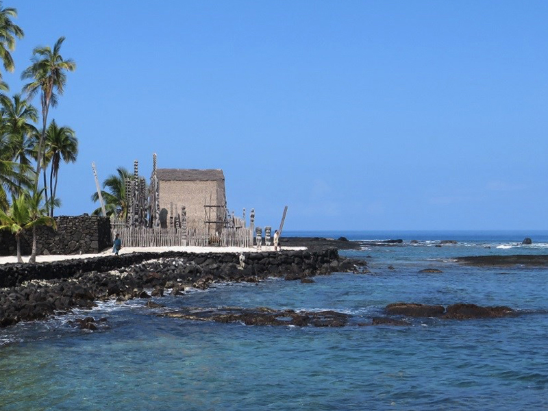 A small historical hut backed by palms sits on a rocky headland with waves in the foreground.
