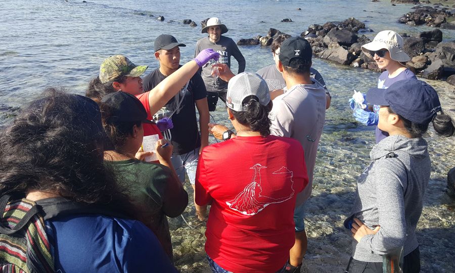 A group of college students on the shore all look to one holding a water sample.