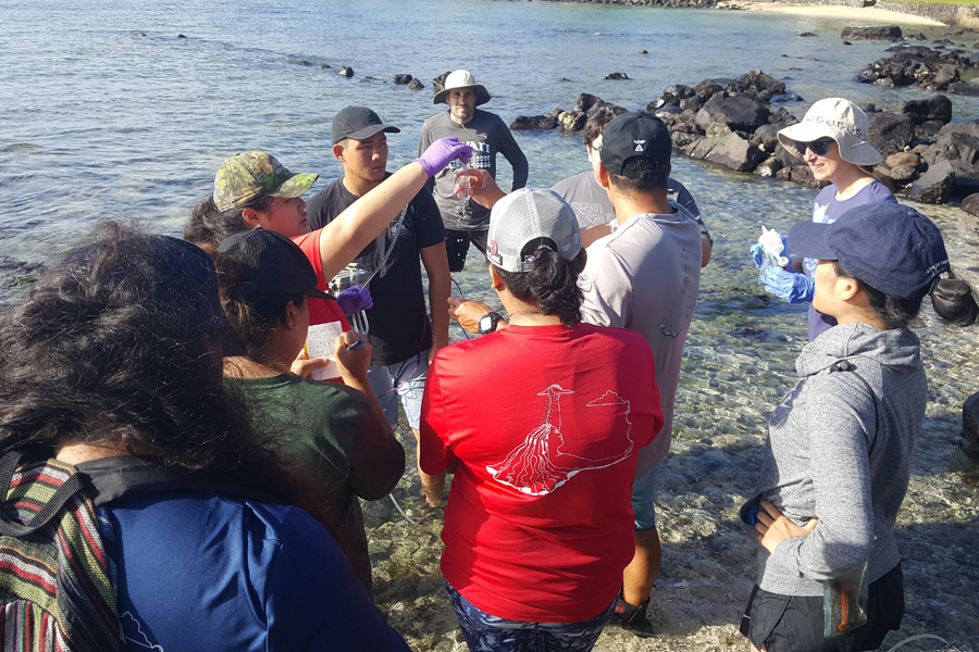 A group of college students on the shore all look to one holding a water sample.