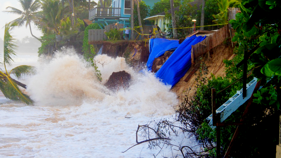 High surf pounds an eroded shoreline, undercutting a bluff beneath houses.