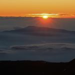 A glowing sun sinks behind a cloudbank with mountains peeking out of the cloud layer in the midground.