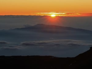 A glowing sun sinks behind a cloudbank with mountains peeking out of the cloud layer in the midground.