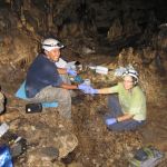 Two students in hardhats smile at the camera while seated on the floor of a cave, with water sampling gear around them.