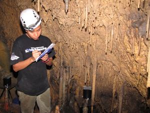 A student recording information stands next to a steeply sloping cave wall covered in stalactites.