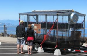 A two-level greenhouse on wheels sits in a parking lot for visitors to look at.