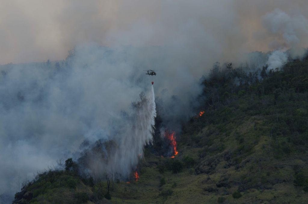 Against a smokey background, a helicopter drops a stream of water on a fire burning up a vegetated ridge
