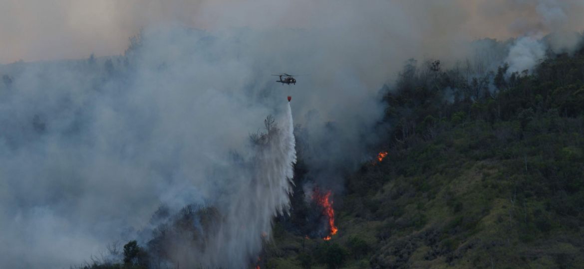 Against a smoky background, a helicopter drops a stream of water on a fire burning up a vegetated ridge