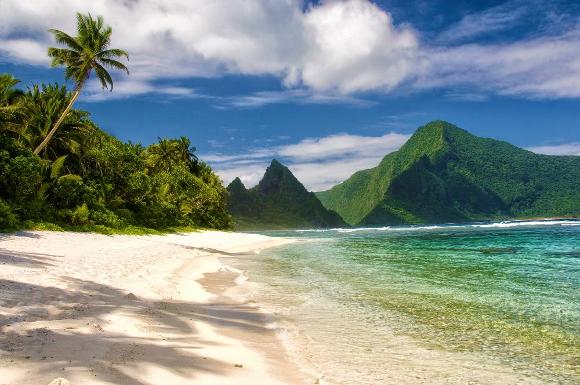 View down a white sandy beach overhung with palms, being lapped by sparkling waters with steep green mountains further along the coast