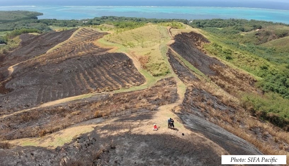 View across a green hill with dark patches of burn scars and blue, tropical ocean in the background.