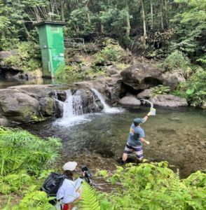 Water flows over a short rocky drop into a pool where a researcher has just sampled the water, while a student sits on the bank with gear