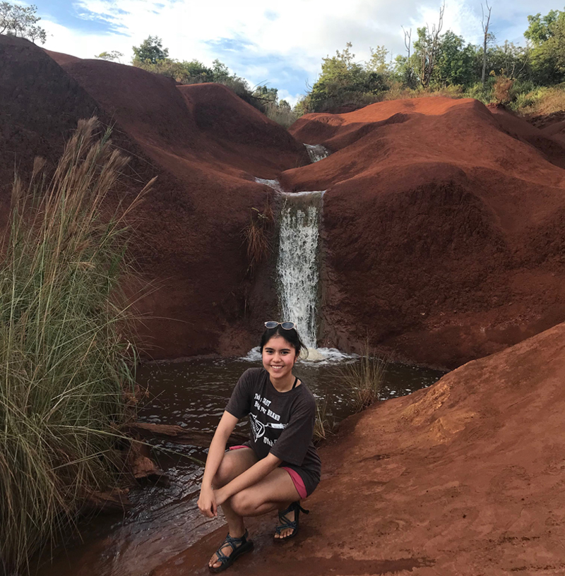 A student crouches on barren red dirt near a cluster of invasive grasses and in front of a running stream