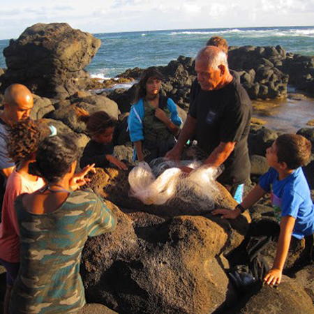 An elder shows students a fishing net at the edge of a rocky pool