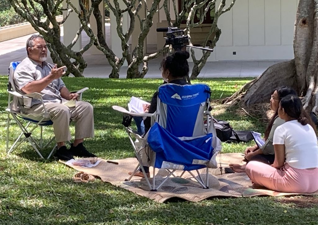 Three young women sit on a woven mat under a shade tree, listening to a middle-aged man gesturing and talking