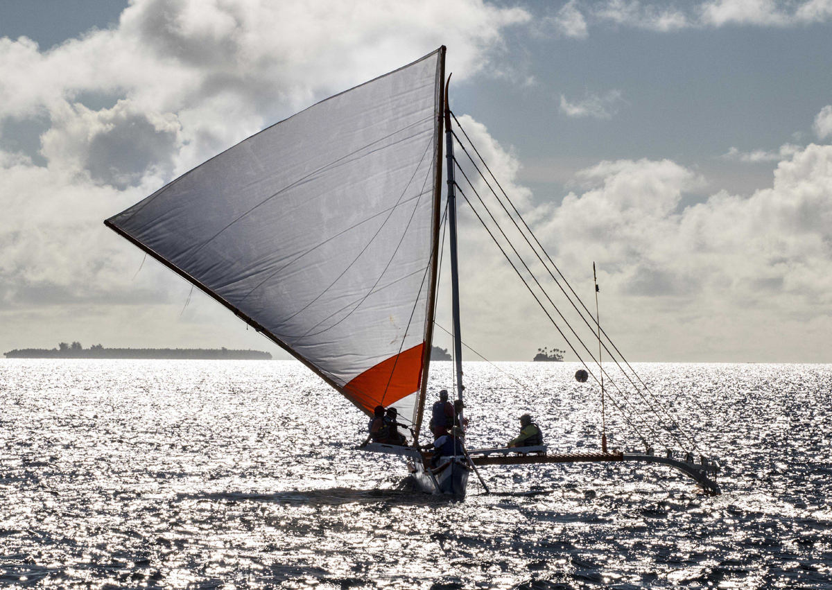 A double-hulled sailing canoe, with sail fully extended for a downwind run, is silhouetted against a sparkling sea.