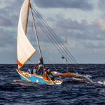 A crew of six in life vests steer a colorful double-hulled sailing canoe on deep blue ocean waters