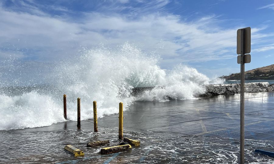 Waves crash over a stone wall and flow across a cemented parking lot, beneath a blue sky