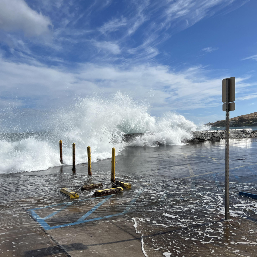 Waves crash over a stone wall and flow across a cemented parking lot, beneath a blue sky