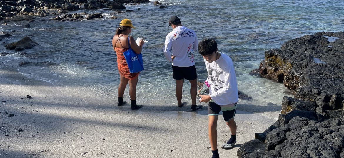 Two students stand recording data at the water’s edge near a black rocky outcropping, while another student walks up the sand.