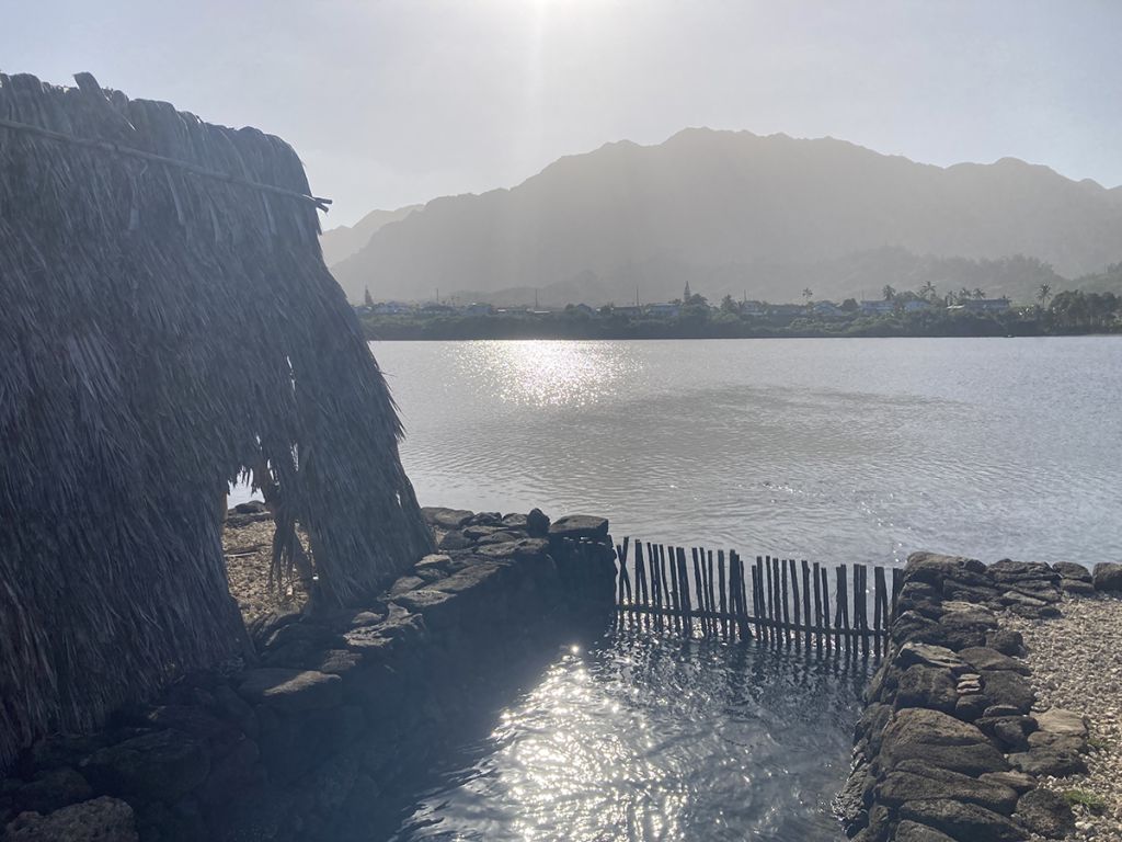 View over a makaha (wooden gate in rock wall) towards open fishpond waters, with a grass shack to the left