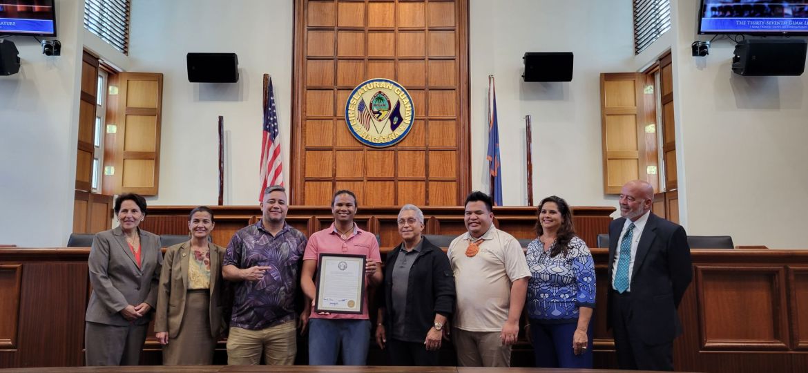 Eight men and women are standing side by side for a picture. A man in the center, wearing a pink shirt, is holding a certificate.