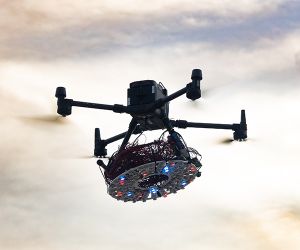Four armed drone flies through the sky with round, heavily wired instrument panel hanging from it