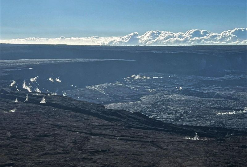 View across barren laval flows, dropping to a sunken caldera floor, with white cumulus clouds on the horizon