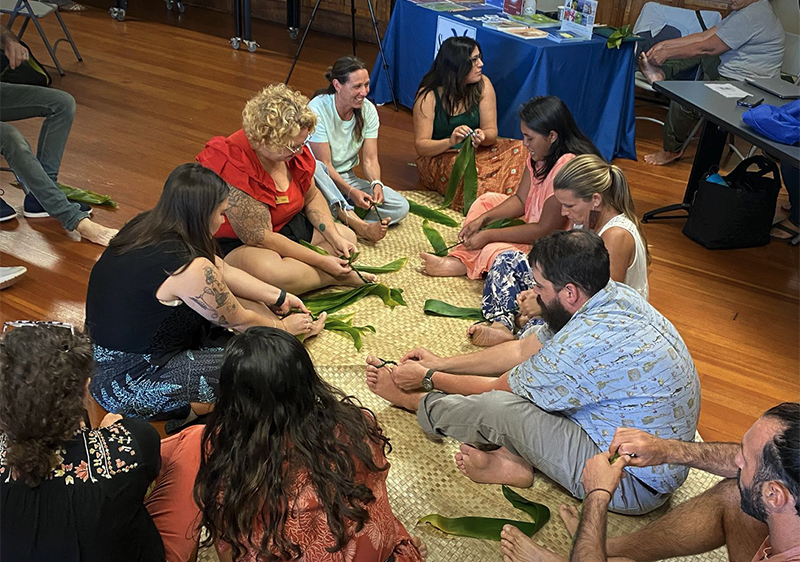 Participants sit around a woven mat, weaving ti leaves together into lei