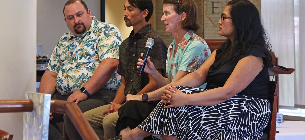 Two men and two women are seated in a row, looking onward. One of the women is holding a microphone.