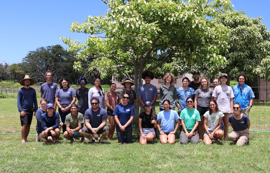 A group of 24 undergraduate and graduate students, and other adults pose for the camera beneath a spreading tree