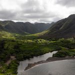 Looking up a broad Hawaiian valley, the foreground shows a bay with an inlet to a medium-sized estuary that disappears into a lush forested valley floor; small waterfalls are visible at the back of the valley in the distance