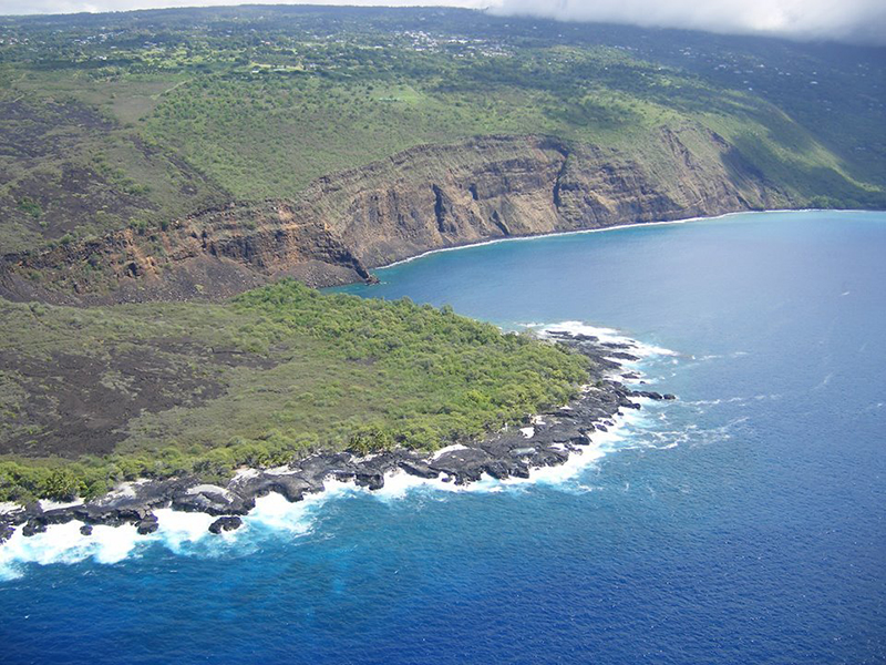 An oblique aerial view of a narrow rocky coastline giving way to steep, layered cliffs defining a broad bay