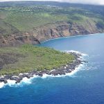 An oblique aerial view of a narrow rocky coastline giving way to steep, layered cliffs defining a broad bay