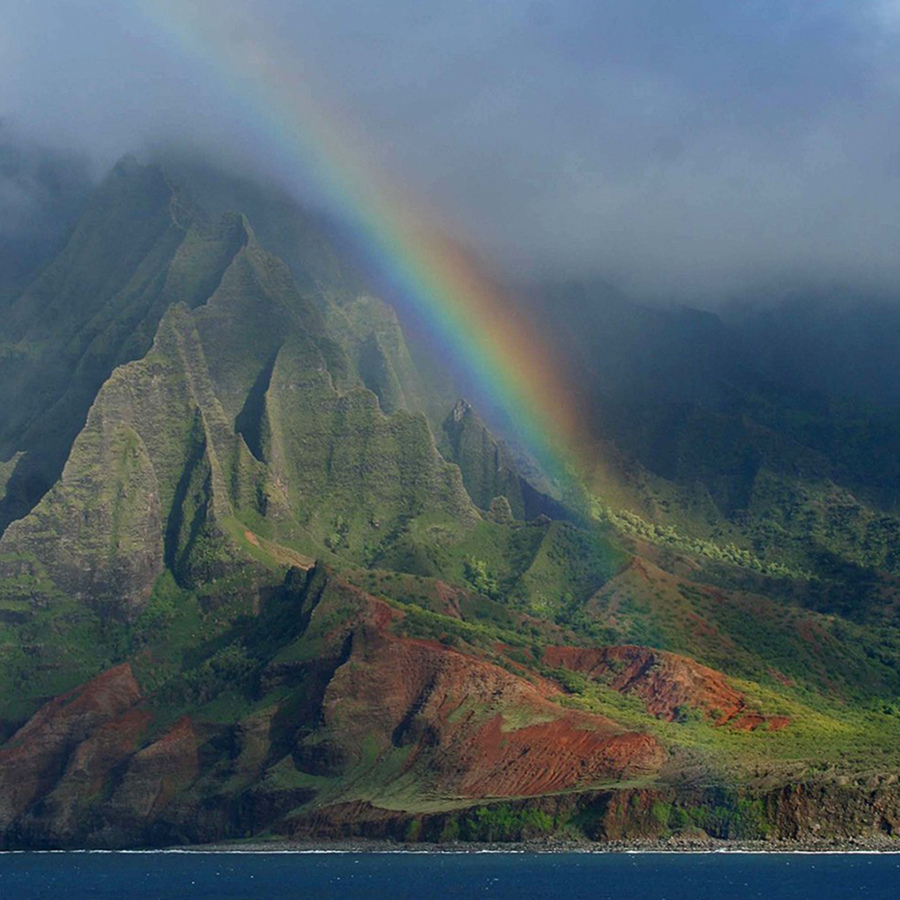 With their tops in the clouds and crossed by a vibrant rainbow, steep, eroded green mountains drop abruptly to a calm ocean coastline