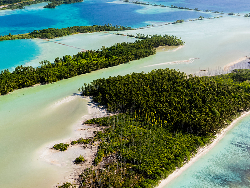 Oblique aerial view of atoll reefs, bright blue and aqua lagoons, and narrow stretches of deep green forests perched on the limited surficial sandbars