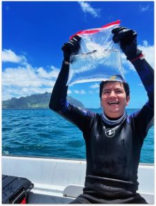 A student seated in a boat and dressed in a wet suit smiles broadly as he holds up a ziplock bag filled with water and fish.