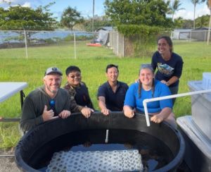 SURF student sends behind four people seated with hands on a large black tub with a white square with dark coral buns on it.