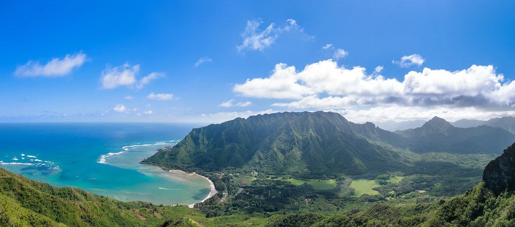 View from high on a ridge looking down over vegetated volcanic terrain to a sea-green bay with blue open ocean beyond