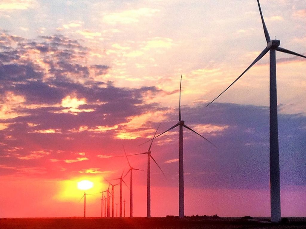 Under a red, cloudy sunset, a line of tall wind turbines stretch into the distance of a flat plain